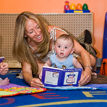 Mom reading a picture book with her baby on the floor of a Kidville classroom