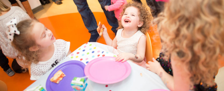 Young girls laughing around a Kidville party table with pizza, pink plates and a polka dot tablecloth during celebration time