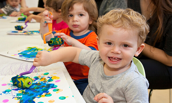 Smiling toddler boy with blue and green paint on his paper at a Kidville messy art table