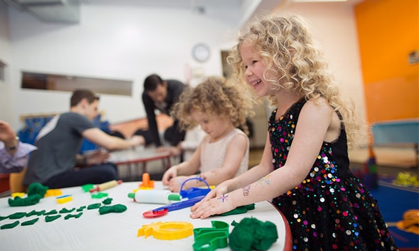Two young girls at a Kidville party table pressing green and yellow playdough shapes during exploration time