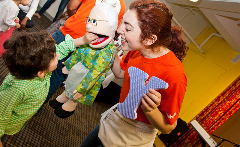 Kidville teacher holding a puppet and a letter cutout while a toddler reaches for it in class at Kidville Upper West Side