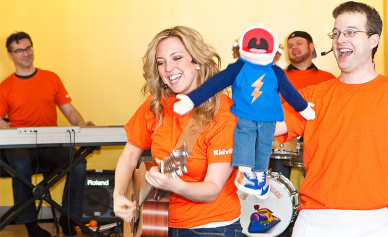 Kidville musicians playing guitar and drums with a puppet during a live toddler music class