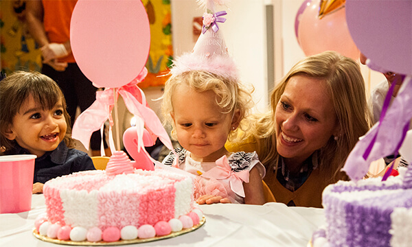 Toddler girl in a pink party hat smiling at her birthday cake as her mom leans in at a Kidville party