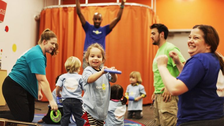 Teachers and young children playing tambourines and dancing together in a Kidville classroom