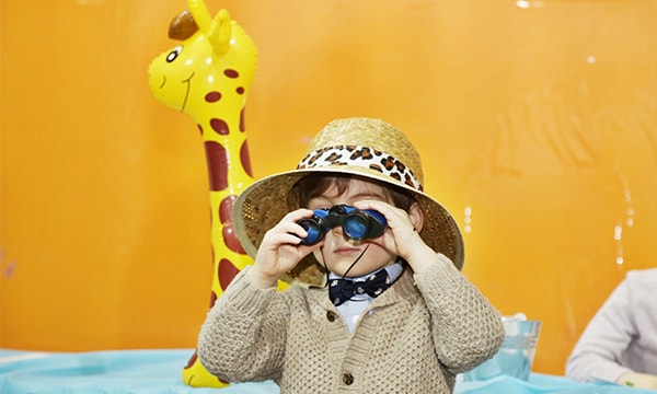 Young Camp Kidville camper in a safari hat looking through toy binoculars in front of an inflatable giraffe on safari theme week