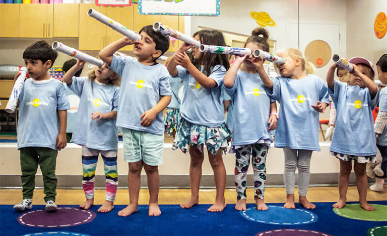 A row of young Camp Kidville campers in matching light blue shirts looking through paper telescopes on space theme week
