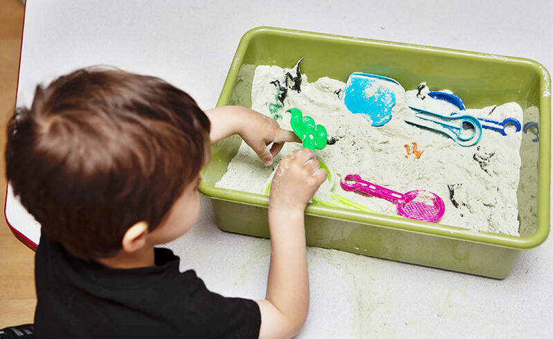 Toddler boy scooping and digging in a green sensory bin filled with sand and small tools