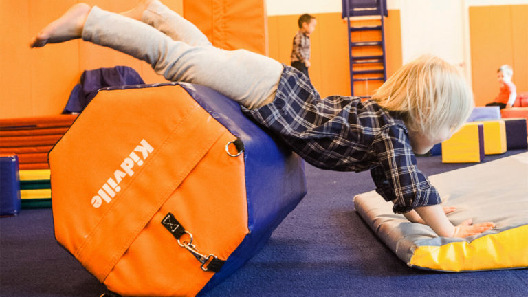 Young child stretched out on an incline mat in the Kidville gym with other children playing behind