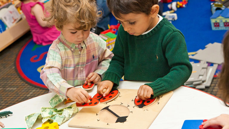 Two young children working on a wooden puzzle together at a low classroom table at Kidville