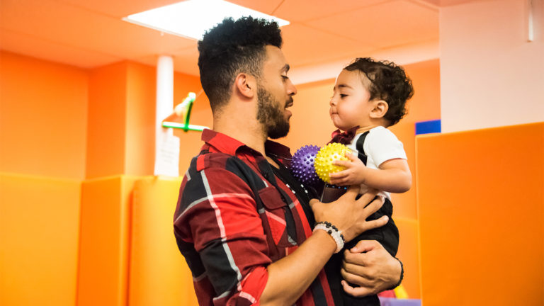 Dad kneeling beside his young child on the mat in the Kidville Upper West Side gym during a parent-and-child class