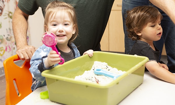 Smiling toddler holding a magnifying tool over a sensory digging bin in class at Kidville Upper West Side