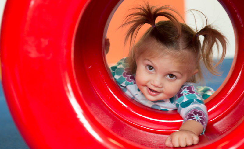 Smiling toddler girl peeking through a red play tunnel in the Kidville gym