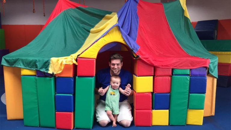 Dad and baby sitting inside a soft block house under a rainbow parachute in the Kidville gym