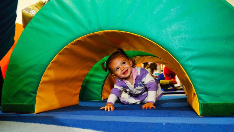 Toddler girl crawling through a green and yellow tunnel on the Kidville gym mats