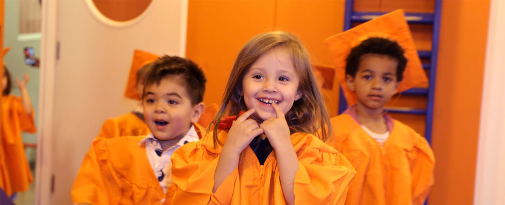 Three young children in orange graduation gowns and caps smiling at Kidville University graduation