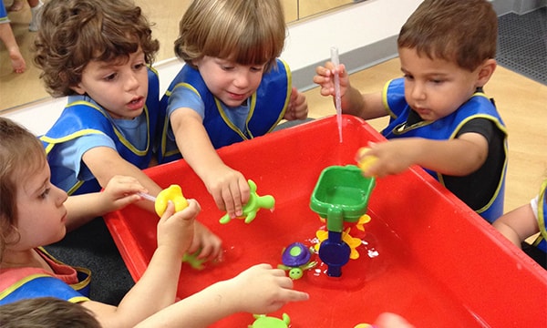 Three toddlers in blue smocks playing together at a red water table at Kidville