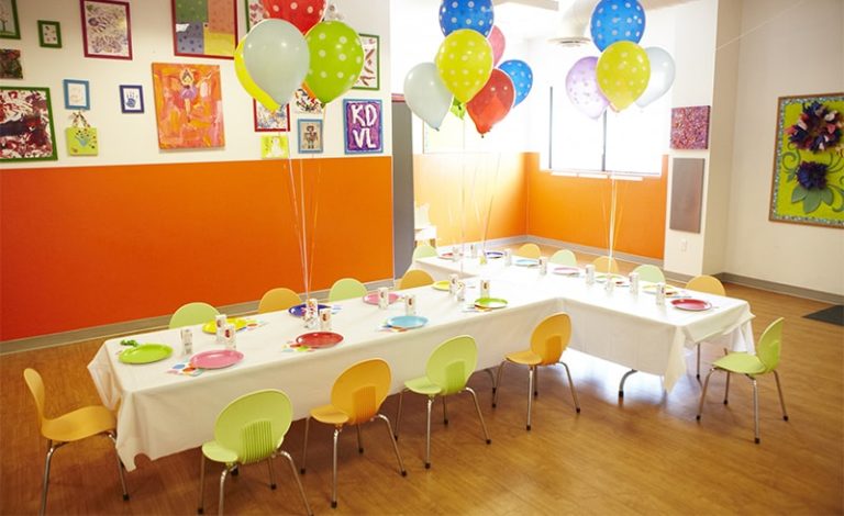 Kidville Upper West Side party room set for a birthday with a long white table, colorful plates, balloon bouquets and green and orange chairs