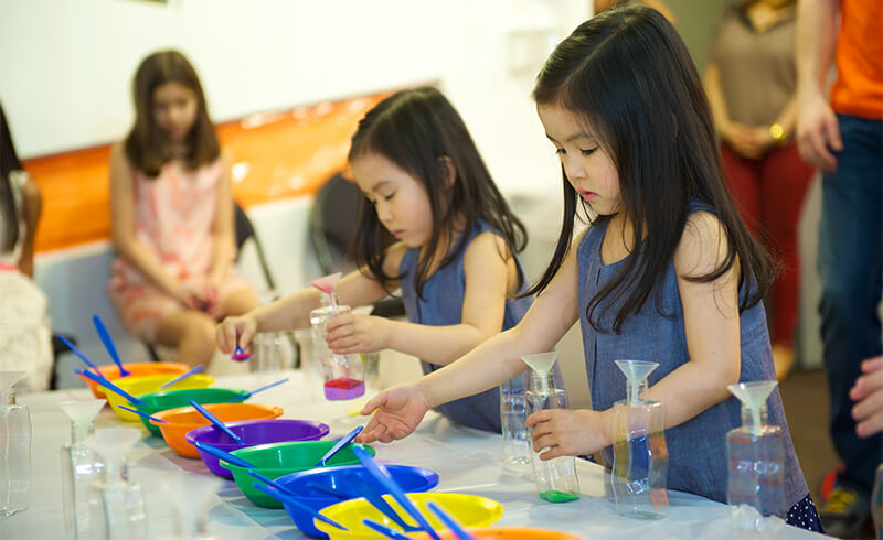 Young girls pouring colored sand into bottles at a Kidville birthday party craft table