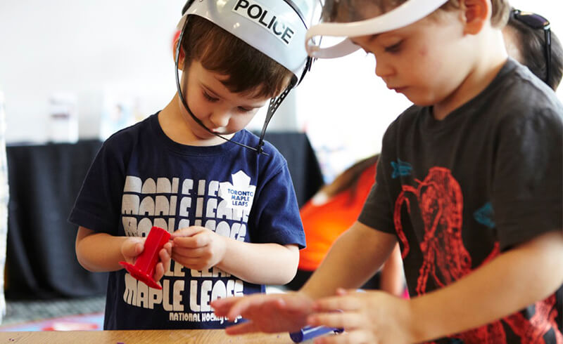 Two preschoolers in play helmets squeezing and shaping red playdough at a table