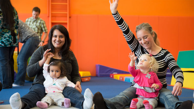 Two moms sitting on the Kidville gym mat with their babies and waving