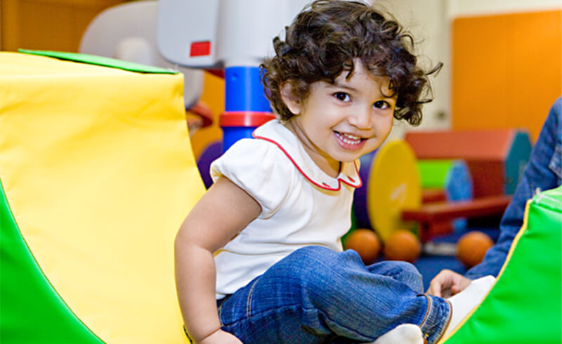 Smiling curly-haired toddler sitting on colorful padded mats in the Kidville gym