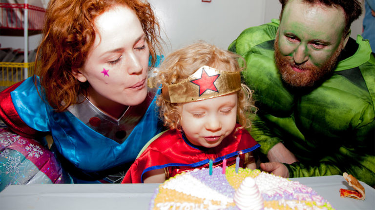 Toddler in a superhero costume and star crown blowing out birthday candles with her costumed parents at Kidville
