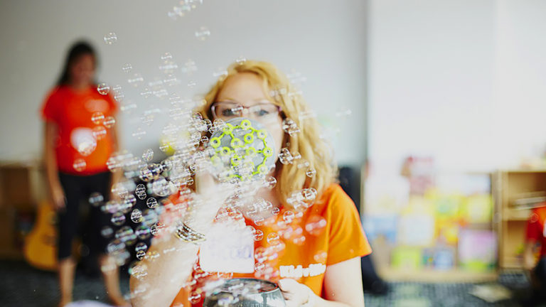 Kidville teacher in an orange shirt blowing bubbles in the classroom